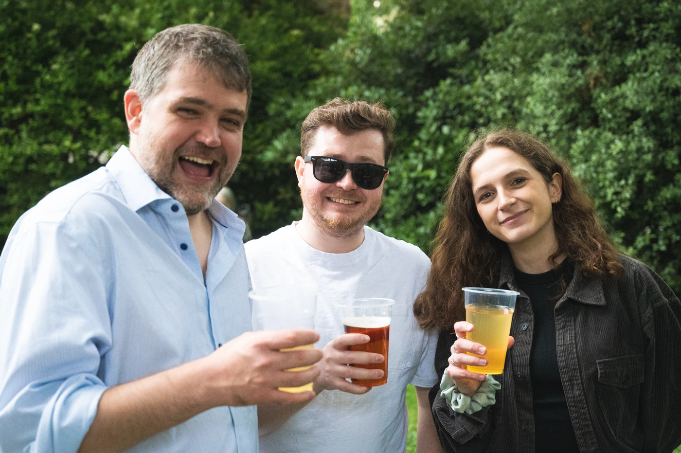 Three people in a garden, each holding a drink in a plastic cup, smiling at the camera