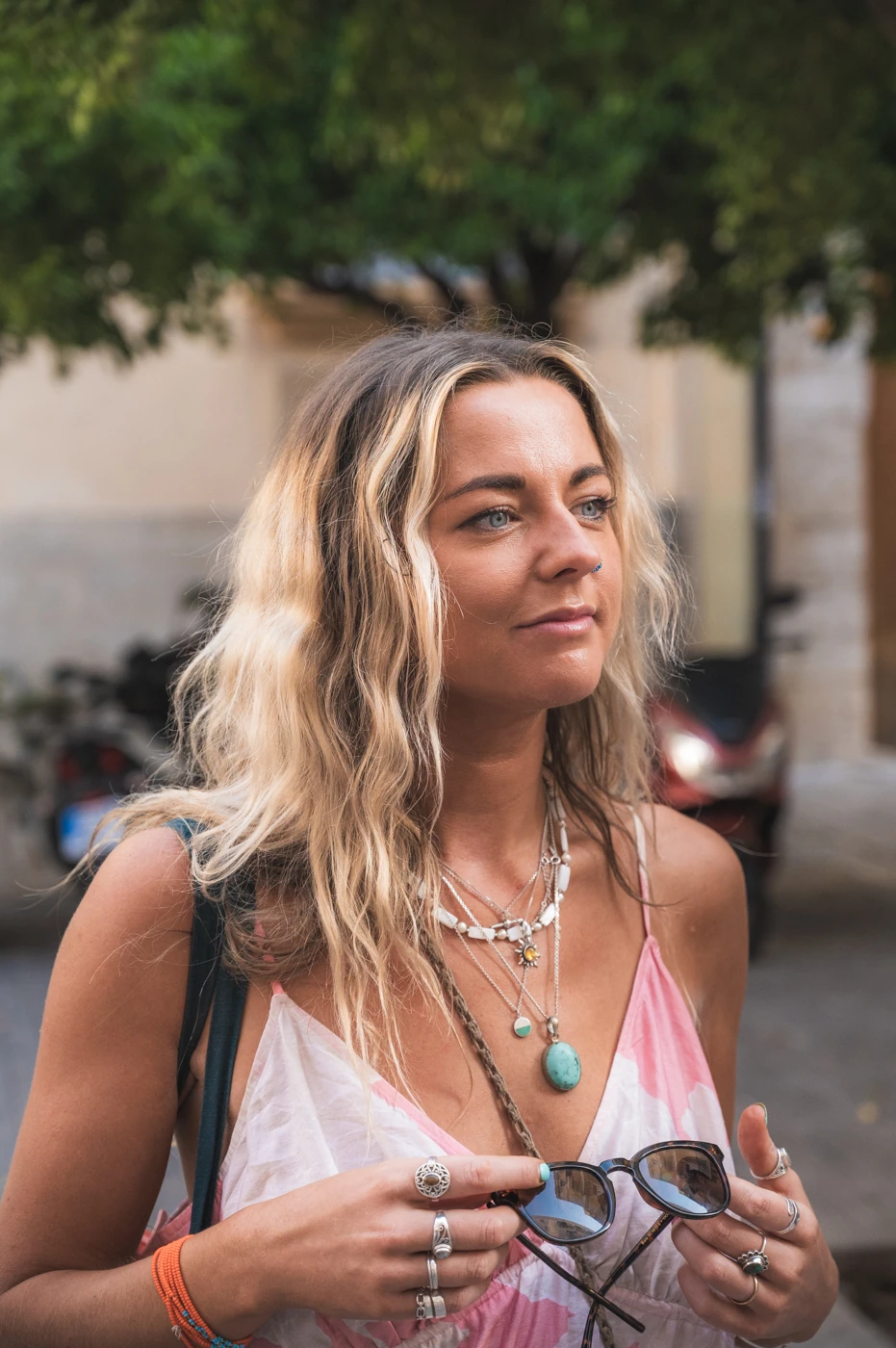 Woman with wavy blond hair wearing layered necklaces and a pink tie-dye top, holding sunglasses outdoors.