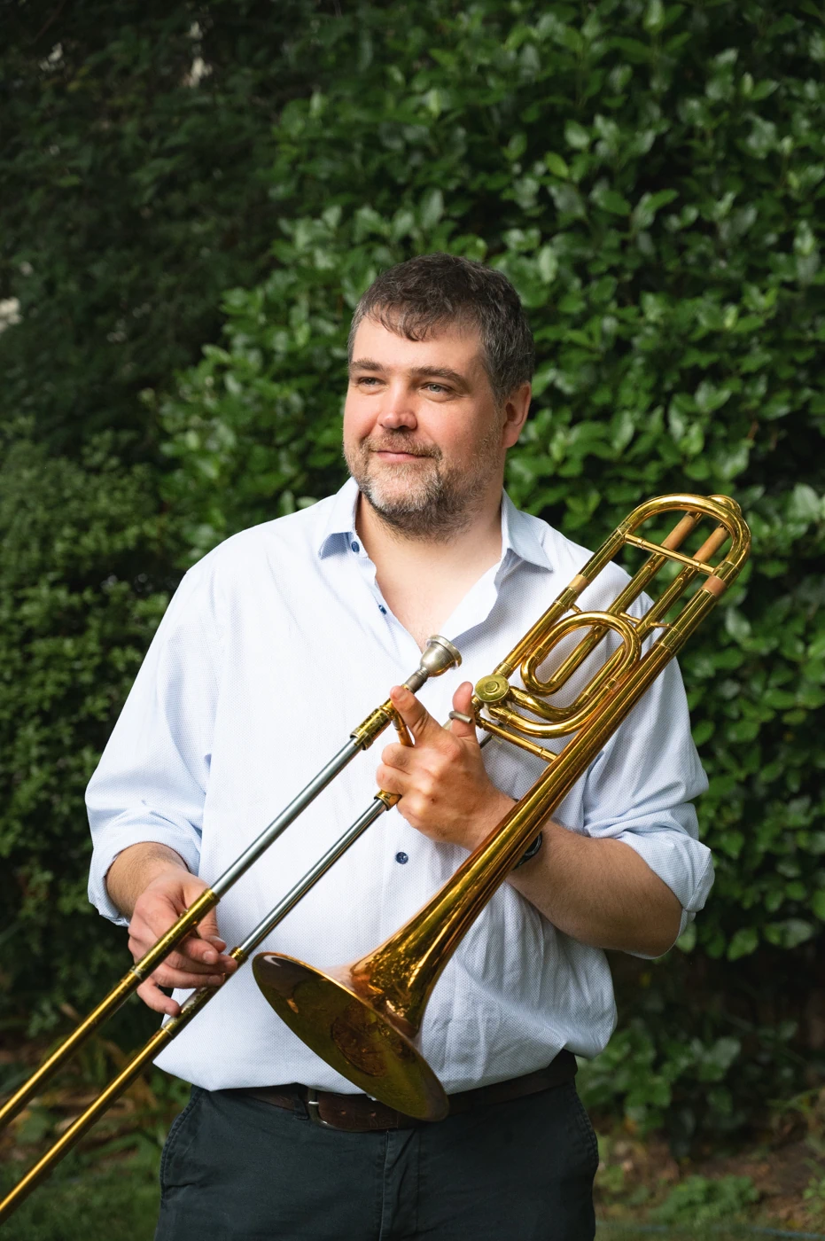 A man standing in front of a hedge, smiling and holding a trombone.