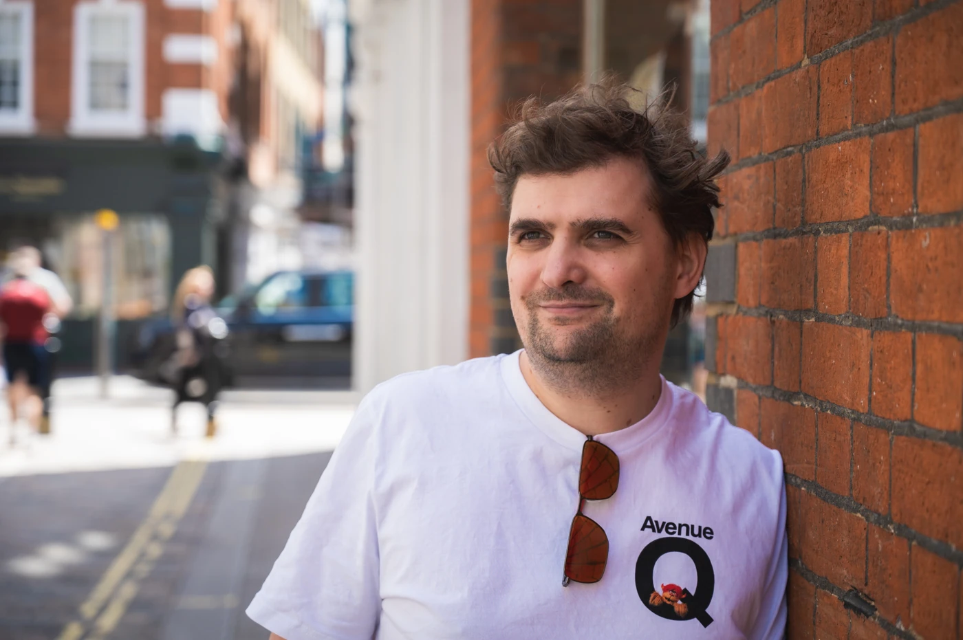 A man smiling, looking away from the camera and leaning against a wall.