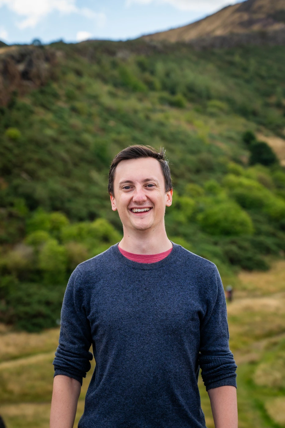 A man outdoors in front of a hill, smiling broadly.