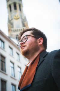 A portrait of a man in glasses facing upwards to reveal the belltower in Bruges