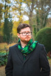 Man with glasses in a green scarf and a dark overcoat, staring into the distance in a garden