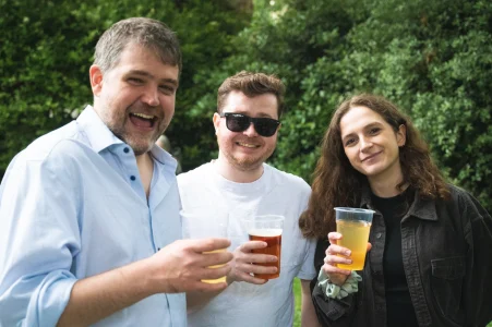 Three people in a garden, each holding a drink in a plastic cup, smiling at the camera