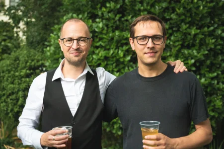 Two men with glasses posing outdoors, each holding a plastic cup and smiling.